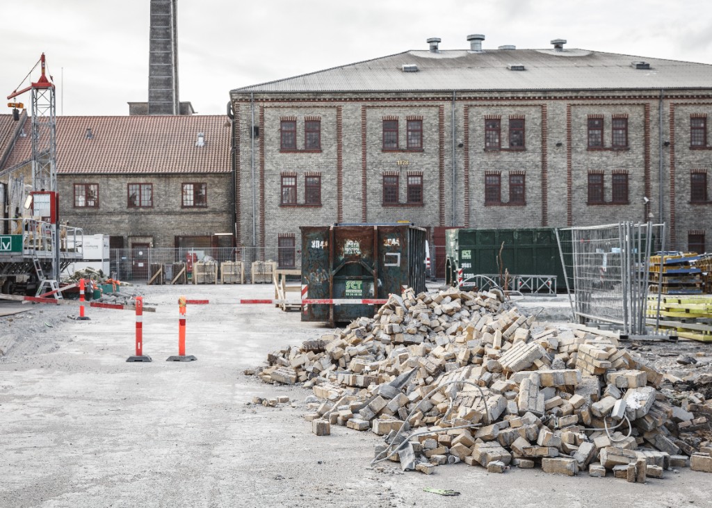 Nedrivningsplads med mursten og containere foran en gammel industribygning.